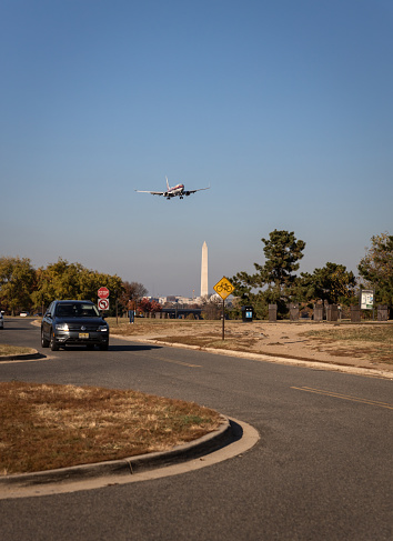 Arlington, United States – November 20, 2023: An aeroplane is seen soaring above a parking lot in the foreground, with the iconic Washington Monument standing at the backdrop