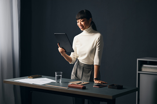 A wide angle view of a smiling Japanese entrepreneur using her tablet while standing in the office.