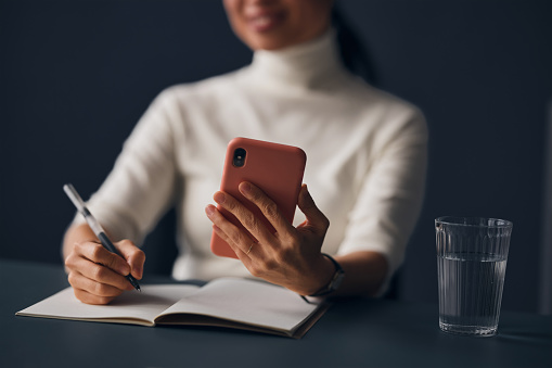A close up view of an unrecognizable Japanese entrepreneur taking some notes while using her smartphone. She is sitting at her office desk.