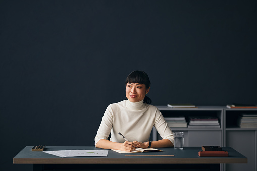 A smiling pensive Japanese entrepreneur contemplating while taking some notes. She is sitting at her office desk.