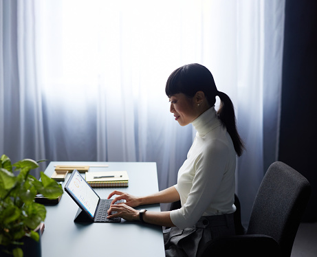 A side view of a smiling Japanese entrepreneur typing on her tablet while sitting at her office desk.