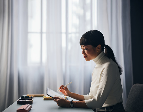 A side view of a smiling Japanese entrepreneur reading something on her tablet while sitting at her office desk.