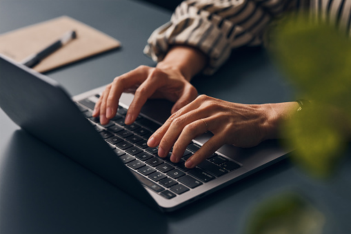 A wide angle view of an unrecognizable Caucasian entrepreneur typing on her laptop while sitting at her office desk. (close up)