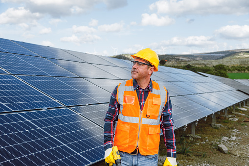 Portrait of a serious mid-adult worker wearing a yellow work helmet and reflective vest. The worker is holding an electrical screwdriver or drill, preparing to install solar panels in a natural sun energy photovoltaic farm power station. The image reflects the commitment and proficiency required for effective solar energy deployment. Ideal for conveying professionalism and dedication to clean energy, this photo is perfect for energy-related promotions, engineering publications, and social media content celebrating advancements in sustainable power. Mid-adult worker, Serious look, Yellow work helmet, Reflective vest, Electrical screwdriver, Drill, Solar panels, Photovoltaic farm, Power station, Expertise, Determination, Clean energy, Energy-related promotions, Engineering publications, Social media, Sustainable power