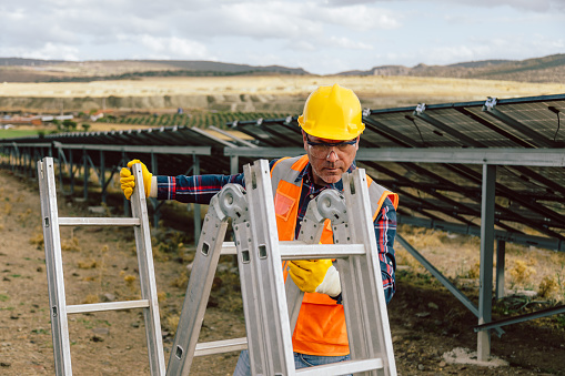Portrait of an engineer or employee middle-aged man holding and preparing the metal folding ladder for work at a natural sun energy photovoltaic farm power station site. The image captures the attention to detail and preparation required for work in the solar energy station area. This effort to ensure the optimal performance of clean and sustainable energy is ideal for energy-related promotions, environmental publications, and social media content celebrating developments in sustainable energy. Solar energy station, Metal folding ladder, Engineer, Employee, Preparation, Sustainable energy, Clean energy, Optimal performance, Energy-related promotions, Environmental publications, Social media, Sustainable energy.