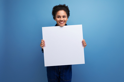 young successful woman with dark skin and black curly hair dressed in blue denim suit holding white blank poster for project showcase.