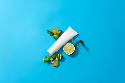 View from above of an unlabeled cosmetic tube with fresh lemon and kumquat displayed against a blue background. Cosmetic mockup with natural extracts for advertising.