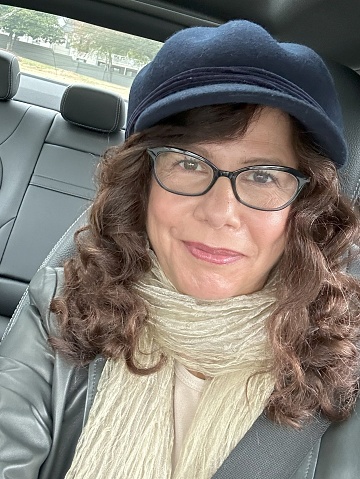 Selfie on mobile device of a senior multiracial woman with glasses and hat on, smiling no teeth and sitting in the drivers seat of her vehicle.