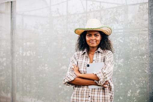 Portrait of a young and successful greenhouse owner wearing a checked shirt and apron standing with crossed arms. A black woman holding a tablet looks at the greenhouse with a smile.