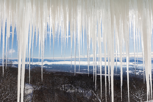 A view through a row of long hanging icicles on a mountain house looking high over a winter landscape.