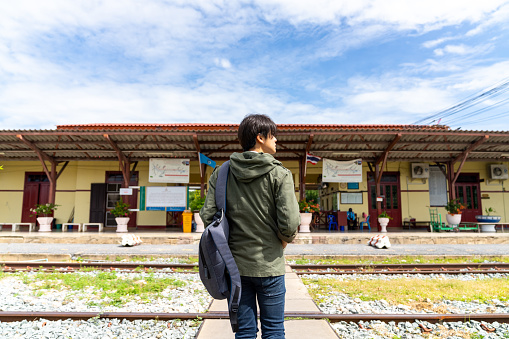 Young Asian man walking on railroad track during waiting for train arrival to station. Handsome guy enjoy outdoor lifestyle travel in Thailand by public transportation on summer holiday vacation.