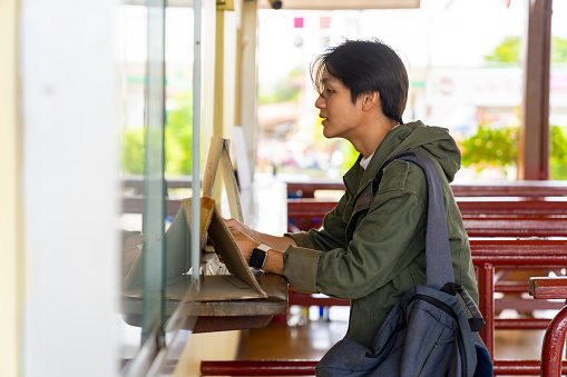 Young Asian man buying train tickets at railway station. Handsome guy enjoy outdoor lifestyle travel local town in Thailand by public transportation on summer holiday vacation.