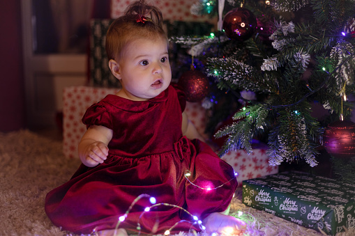 An adorable confused baby girl is sitting on a white carpet in front of a Christmas tree and presents.