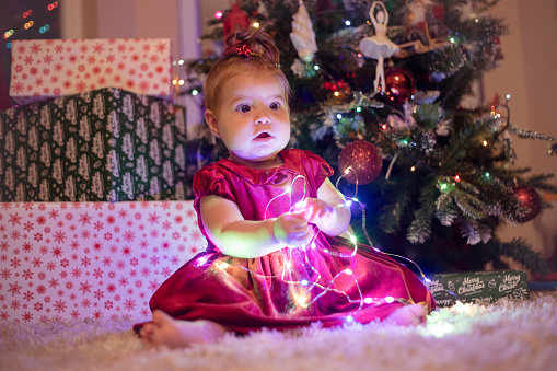 The adorable baby girl is sitting on the white carpet in front of the Christmas tree and playing with LED string lights garland. The child is playing with Christmas decoration .