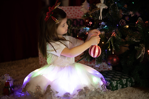 Adorable little girl in a glowing dress is sitting on the white rug in front of the Christmas tree and putting the red ornament on.