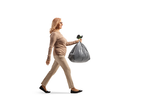 Full length profile shot of a middle aged woman carrying a plastic waste bag isolated on white background