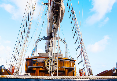 Old wooden sailing ship, closeup. Wooden steering wheel, ropes, rope ladder and bronze bell.
