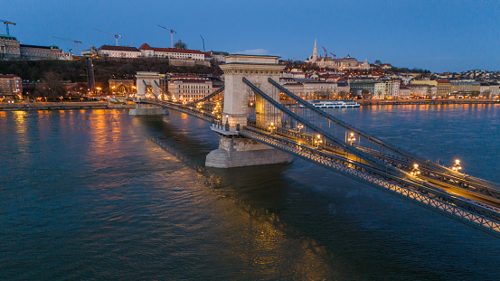 In the Gentle Dusk of Hungary,a High Angle Drone Captures the Ethereal Beauty of Budapest. The Szechenyi Chain Bridge,Adorned with Lights,Elegantly Spans the Danube River. The Scene Unveils the City's Transition From Day to Night