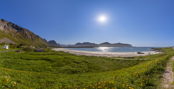 Wide-angle panorama showcasing the radiant sun over Jusnesvika Bay, with lush greenery, yellow wildflowers, white sandy Rambergstranda beach, and people basking in the sun, flanked by Lofoten's rugged mountains