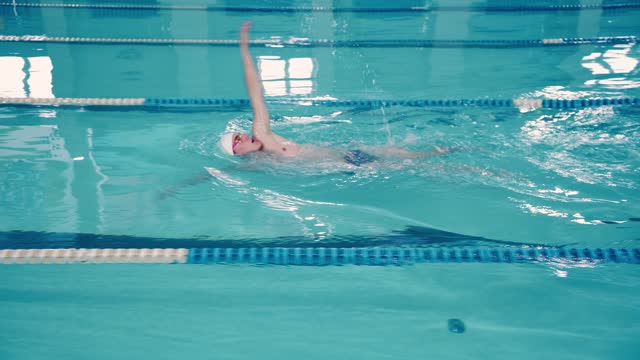 A young male athlete swimmer swims underwater in swimming pool, emerges and continues to swim in the backstroke style.
