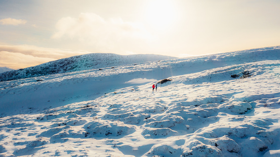 Drone panoramic photo of man in red and woman in blue down jackets contemplating hiking trip at the beautiful winter mountain landscape with a view of the fjord and beautiful lake during snowy blizzard and bright scenic sunset in the western fjords of Scandinavia