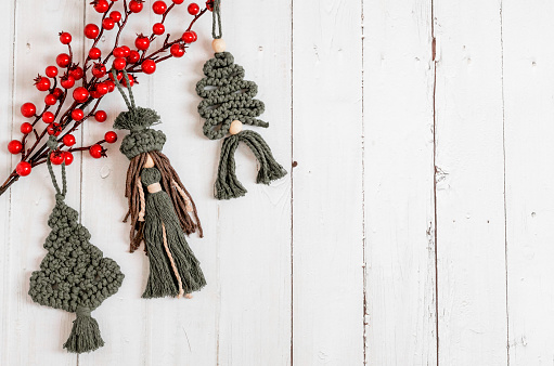 Three hand-knitted woolen Christmas tree toys - a spruce tree and a doll with a branch of red berries lie on the left on a white wooden table with copy space on the right, flat lay close-up.