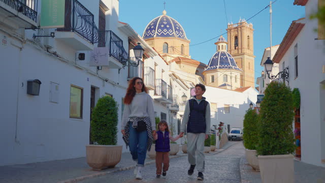 Family walking along a Spanish street in a small village with white houses. Spanish city of Altea in the city center on the mountain church with a blue roof. Tourists sightseeing