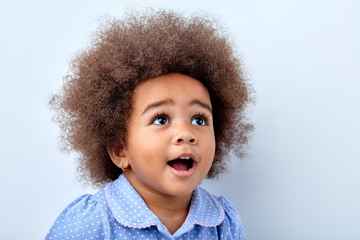 close-up portrait of cute surprised african child girl in blue t-shirt looking up at something interesting, kid girl having curly fluffy hair, smiling happily, expressing sincere genuine emotions