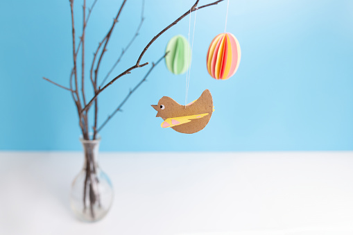a vase filled with Easter decorations on top of a table, a pastel paper bird craft hanging on a tree, soft focus, step by step instruction, DIY, spring craft activity for kids .