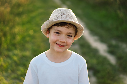 Portrait of a smiling little boy in a white T-shirt and hat playing outdoors on the field at sunset. Happy child, lifestyle. Products for children.