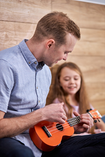 Dad teaching guitar and ukulele to his daughter sitting on bed at home.girl learning guitar with father.Ukulele class at home. free time at weekends. copy space. focus on male playing ukulele