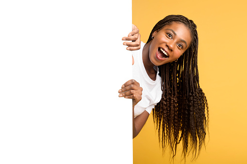 Funny astonished african woman peeps out from behind empty white banner, hiding behind huge paper poster, girl with pigtails surprised with deal, advertising, mock up, copy space, isolated on yellow
