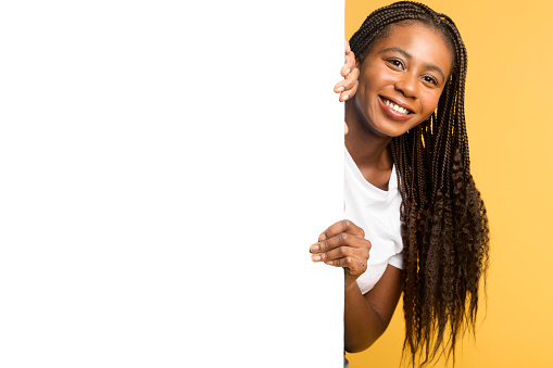 Advertising concept. Smiling attractive black woman peeking out from behind white empty paper poster, presenting newelty, charming african-american girl with blank banner isolated on yellow, copyspace