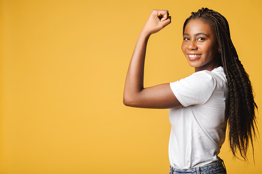 Female power. Young african-american woman showing bicep and looking at the camera isolated on yellow, portrait of smiling strong black determined woman