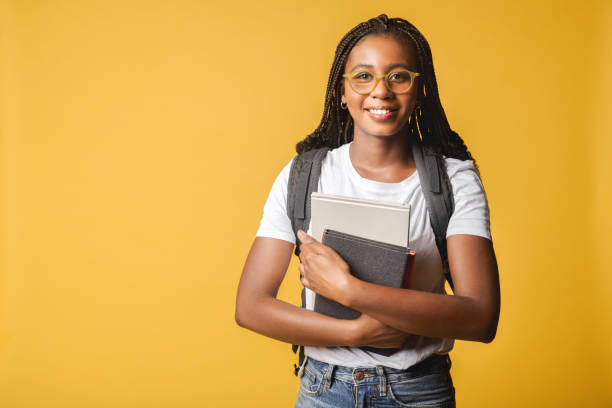 bonita y atractiva joven estudiante de 20 años con camiseta blanca básica y gafas de pie con mochila y llevando libros aislados en amarillo - back to school young women cheerful happiness fotografías e imágenes de stock