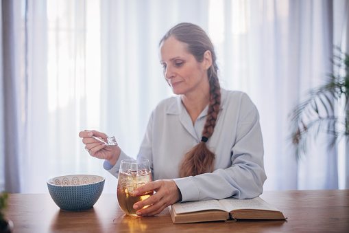 A middle-aged Caucasian woman carefully removes ice from her glass to avoid tooth sensitivity, seated at home with a book.