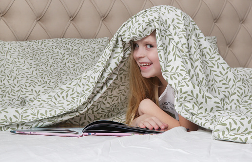 Adorable little preschool girl holding hiding under blanket with book in the bedroom, smilling and looking at camera