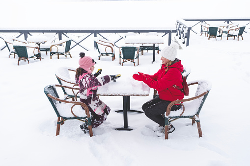 An empty cafe in the park in winter. A young woman and a girl of 8 years old are sitting on chairs at a table outside in a snowy park.