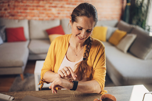 A smiling middle-aged Caucasian woman engages with her smartwatch, seamlessly integrating technology into her work routine at a cozy home office.