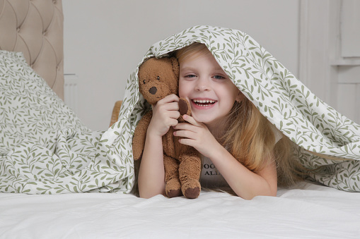Adorable little preschool girl holding her teddy bear and hiding under blanket in the bedroom