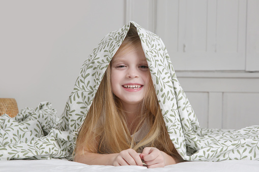 Adorable little preschool girl hiding under blanket, smilling and looking at camera