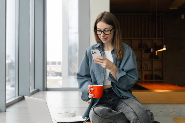 istock Young female student holding a cup and using a smartphone near the window. 1884958192
