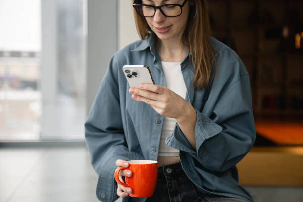 istock A woman is using a smartphone and holding a cup while sitting by the window. 1884961950