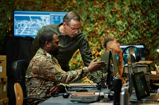 Young African American military officer in camouflage uniform pointing at computer screen while explaining graphic data to colleague
