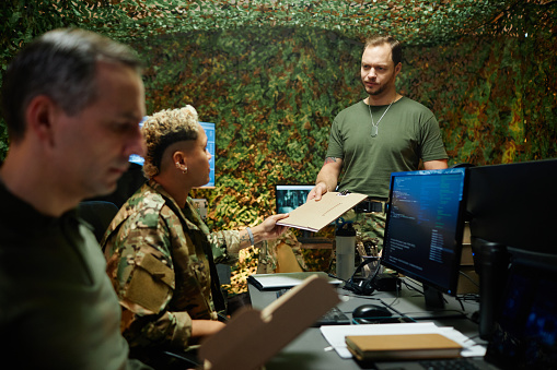 Young male officer in t-shirt passing folder with secret documents to multiethnic female colleague in camouflage uniform