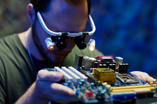 Focus on young repairman with special eyewear with magnifying lens looking at part of processor with microchip and other details