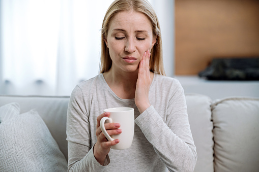 Portrait of sad woman sitting on sofa, holding cup, feeling pain while drinking hot tea or cold beverage. Sensitive teeth. Dental and oral care for problem enamel