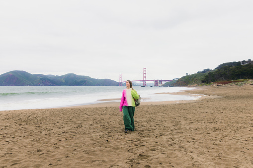 Front view of woman with long hair, in colorful knitted sweater walking at the scenic beach and enjoying music from headphones with background view of red Golden Fate Bridge in San Francisco city, the United States
