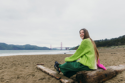Side view of smiling woman with long hair, in colourful knitted sweater sitting at the scenic beach with background view of red Golden Fate Bridge in San Francisco city, the United States