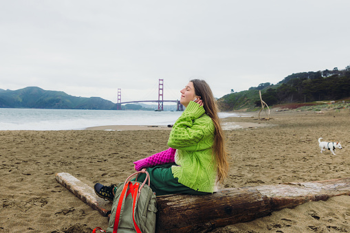 Side view of smiling woman with long hair, in colorful knitted sweater sitting at the scenic beach and enjoying music from headphones with background view of red Golden Fate Bridge in San Francisco city, the United States